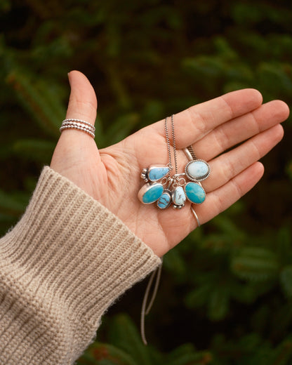 Hand holding three silver necklaces with turquoise stones against a blurred green background